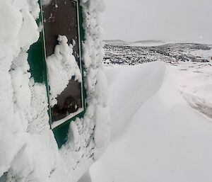 A building in a snow covered landscape with a snow drift just outside.  A window from the building has been partially cleared of snow.