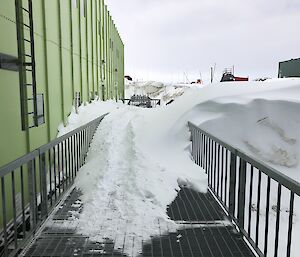 A walkway next to a green building with deep snow drifting over the handrails