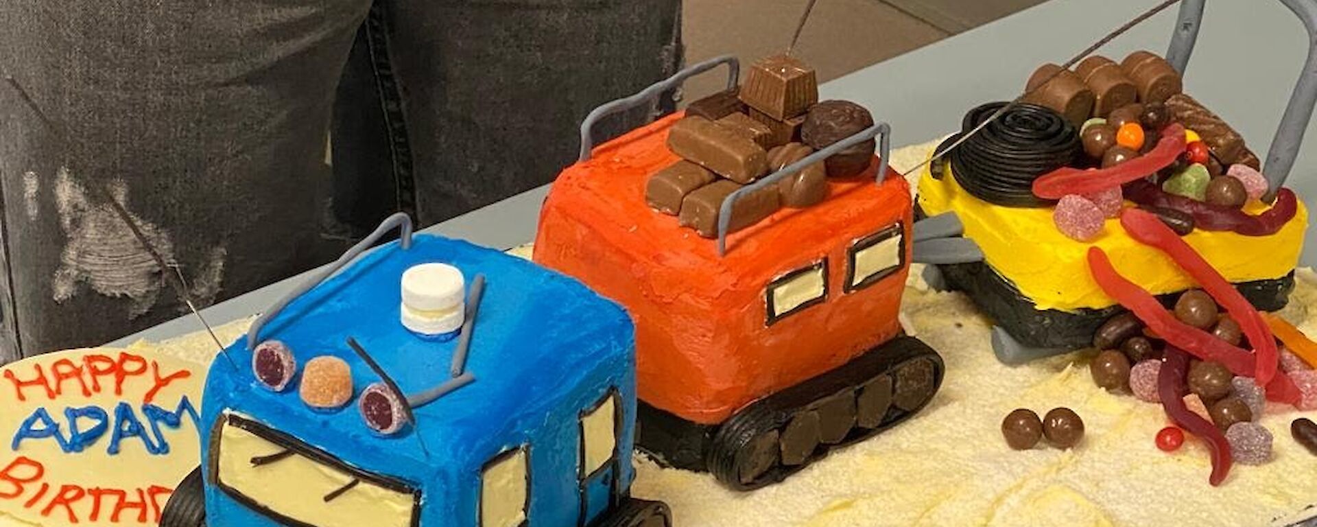 Birthday cake in the shape of a blue and orange Hagglund over snow vehicle towing a yellow sled.