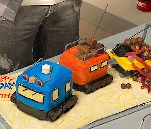 Birthday cake in the shape of a blue and orange Hagglund over snow vehicle towing a yellow sled.