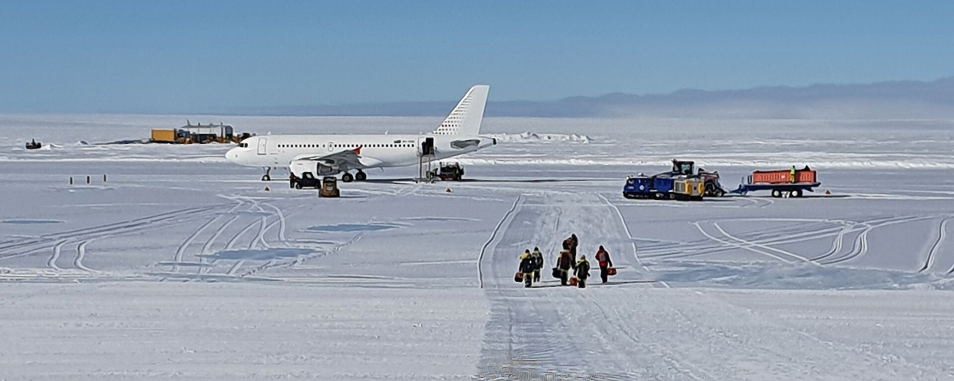 An ice runway with a plane parked at the end and expeditioners with bags walking towards camera