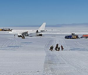 An ice runway with a plane parked at the end and expeditioners with bags walking towards camera