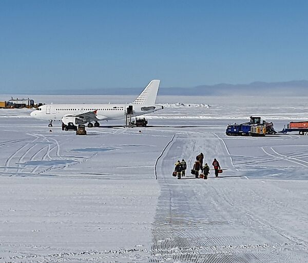 An ice runway with a plane parked at the end and expeditioners with bags walking towards camera