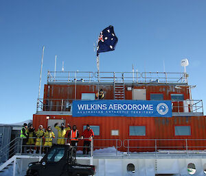 A red building in the middle of a snowy landscape, with the Australian flag flying above.  Serveral expeditioners are gathered on the walkway up to the building.  A sign reads Wilkins Aerdrome.