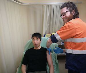 An expeditioner sits on a hospital bed sucking a lollipop with his sleeve rolled up,  whilst a doctor administers a vaccine