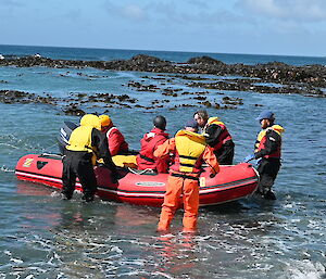 A red dinghy close to shore with people in life jackets pulling it towards shore