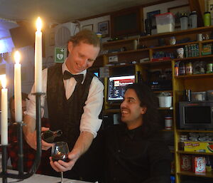 A man pouring a glass of red wine for a dinner guest by candle light