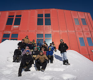 Group of expeditioners sitting on a sloped blizz tail in front of a red building