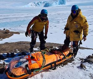 Two expeditioners standing next to a stretcher on the ground.  Icebergs and ice behind them.