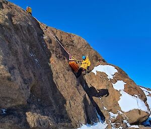 The stretcher attendant and stretcher being raised up the rock face on ropes.