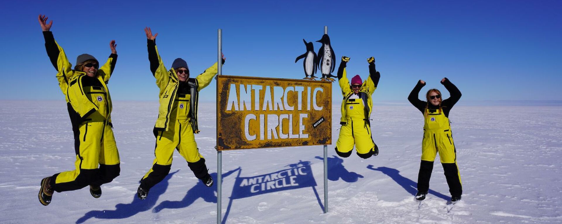 Four woman jumping with excitement next to the Antarctic Circle sign