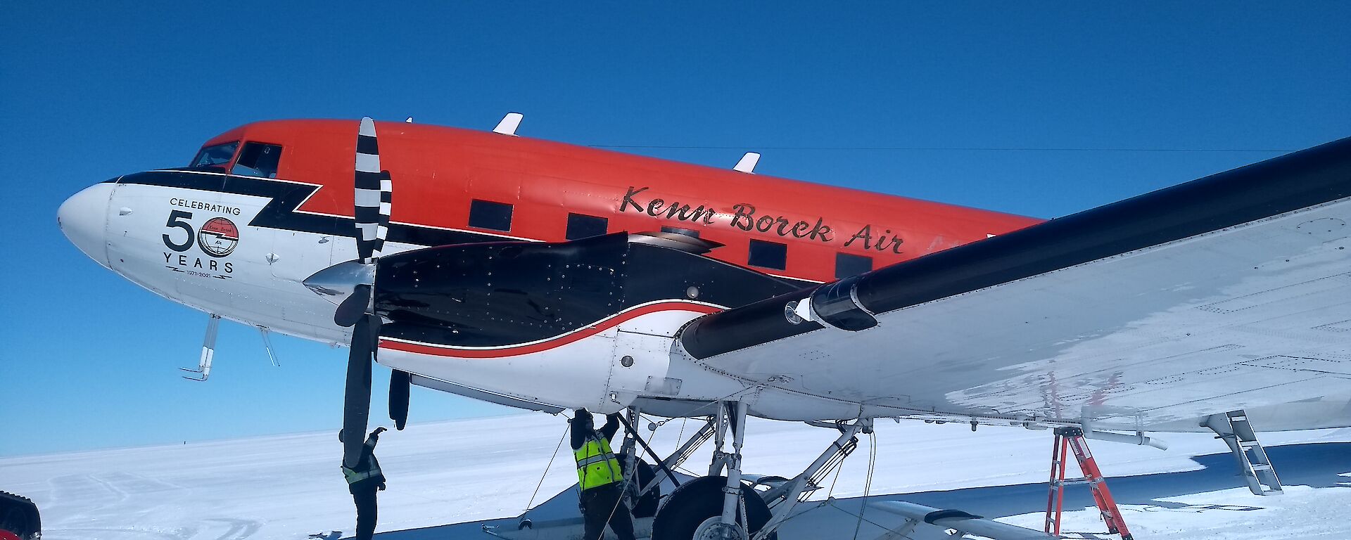 A red DC-3 Basler parked on the snow with a person working underneath it