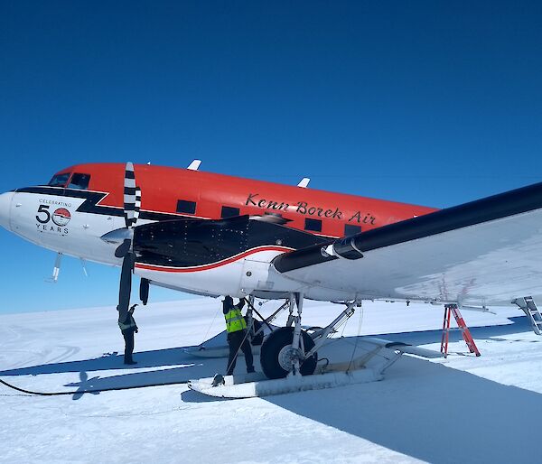 A red DC-3 Basler parked on the snow with a person working underneath it