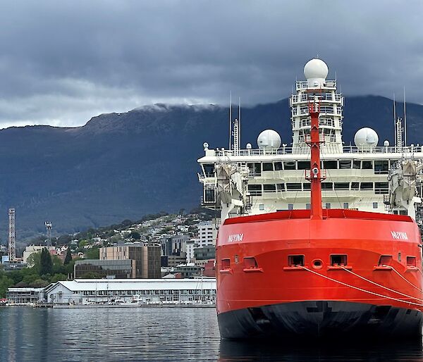 A large red ship tied to a dock with buildings and a mountain behind.  Clouds cover the top of the mountain.