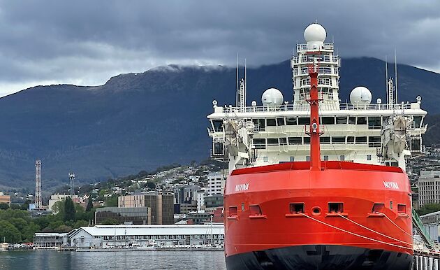 A large red ship tied to a dock with buildings and a mountain behind.  Clouds cover the top of the mountain.