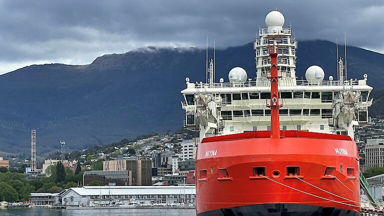 A large red ship tied to a dock with buildings and a mountain behind.  Clouds cover the top of the mountain.