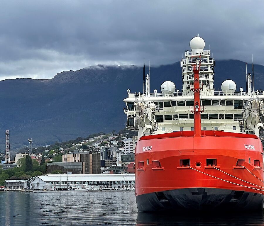 A large red ship tied to a dock with buildings and a mountain behind.  Clouds cover the top of the mountain.