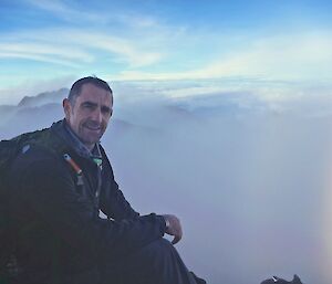 A man sits on top of a mountain with clouds behind him