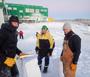 Three men in beanies building an igloo with the green living quarters building in the background.