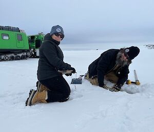 Two people kneeling on the ice with a green Hagglunds in the background