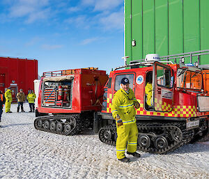 A red Hägglunds vehicle equipped for firefighting with hoses, extinguishers and other equipment. One member of the fire team stands in front by the vehicle door, others stand in the background