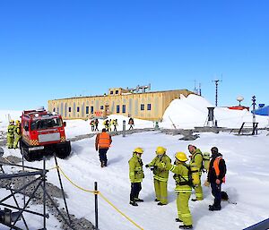 A team of expeditioners standing in the snow in firefighting gear. surrounding by fire vehicle and buildings at Casey Station