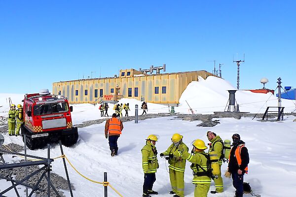 A team of expeditioners standing in the snow in firefighting gear. surrounding by fire vehicle and buildings at Casey Station