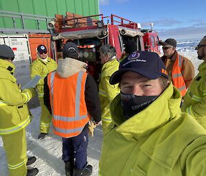 Expeditioners inspecting the pump controls on the back of the fire vehicle. Fire chief in the foreground smiling behind his balaclava