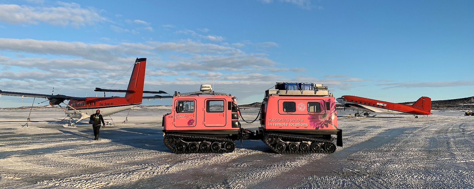 A man walking next to 2 planes and a pink Hagglunds on the sea ice