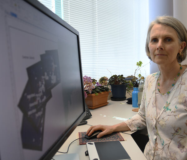 A woman sits at a desk, looking to camera.  A series of Landsat images are on her screen