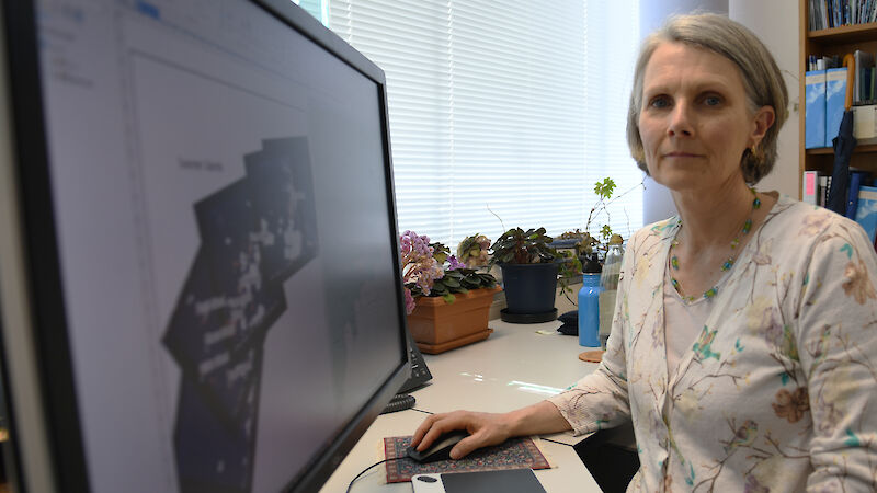 A woman sits at a desk, looking to camera.  A series of Landsat images are on her screen