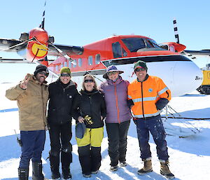 A group of five people standing smiling to camera in front of a twin otter plane on the sea ice