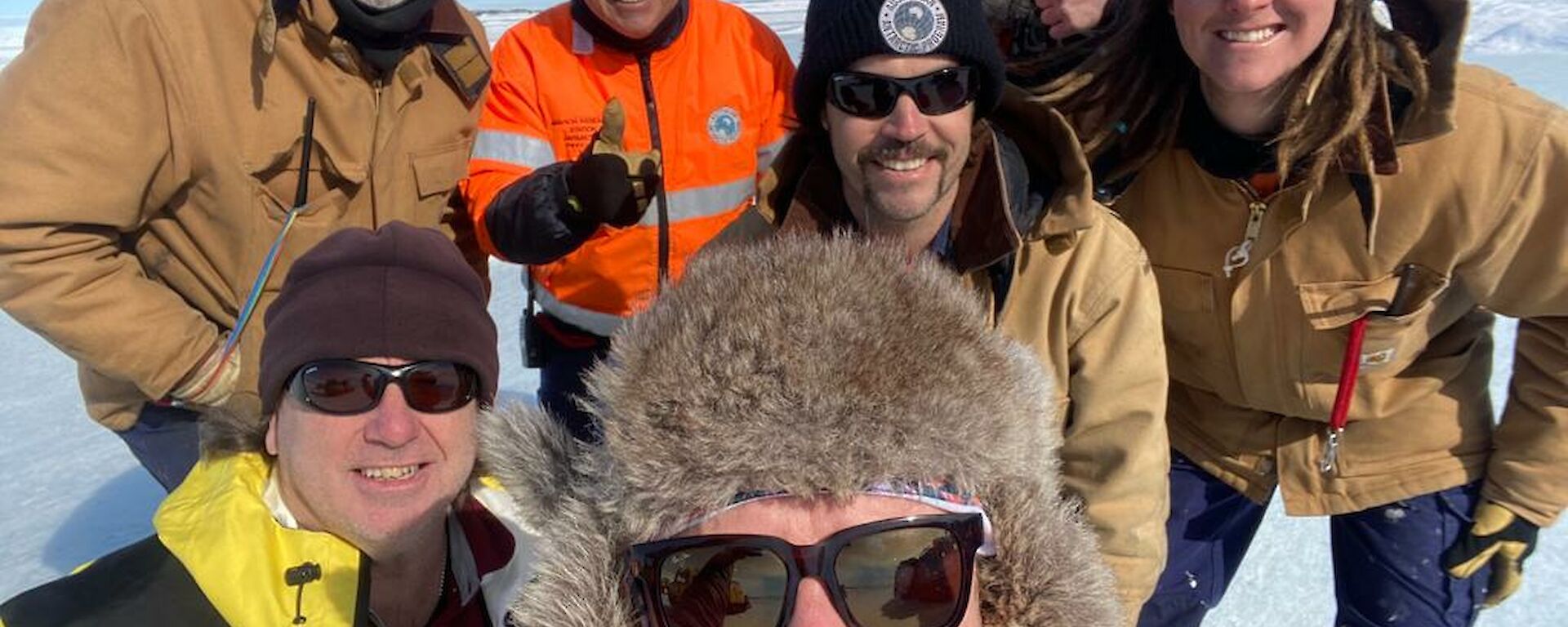 A group selfie of expeditioners on the ice, wearing fur hats, beanies, sunnies and other warm weather clothing