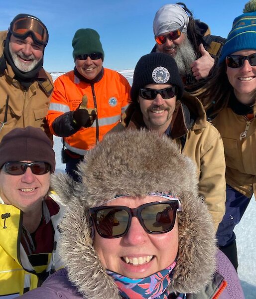 A group selfie of expeditioners on the ice, wearing fur hats, beanies, sunnies and other warm weather clothing