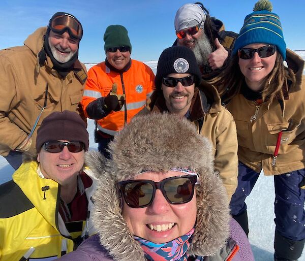 A group selfie of expeditioners on the ice, wearing fur hats, beanies, sunnies and other warm weather clothing