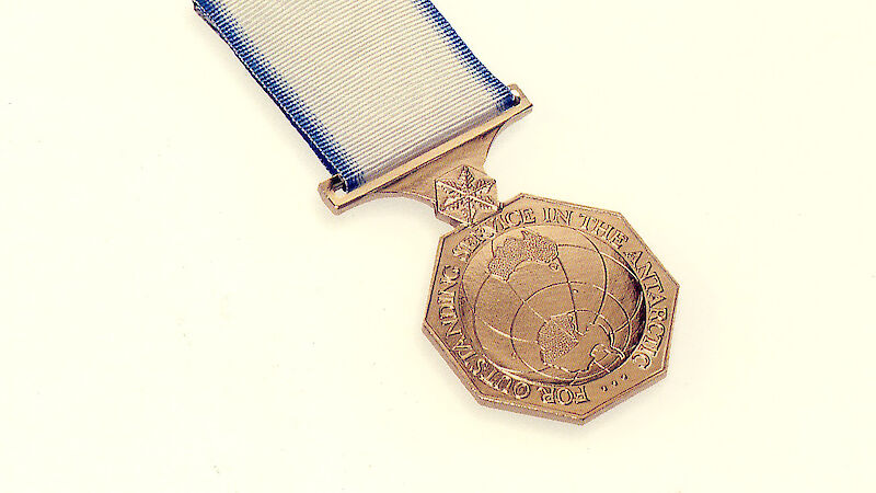 A Hexagonal medal with a blue and white ribbon.  Wording on the medal reads "For outstanding service in the Antarctic"