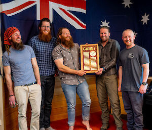 Five members of the outgoing Casey team standing before an Australian flag. Two of them hold up a plaque engraved with the names of all their team members