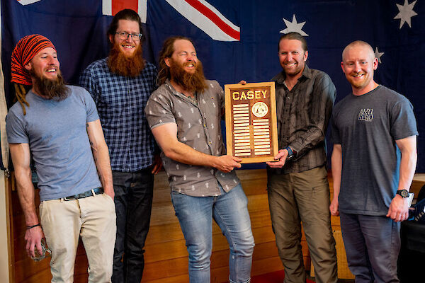 Five members of the outgoing Casey team standing before an Australian flag. Two of them hold up a plaque engraved with the names of all their team members