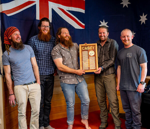 Five members of the outgoing Casey team standing before an Australian flag. Two of them hold up a plaque engraved with the names of all their team members