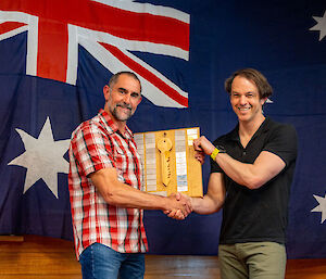 The old and new station leaders posed in front of the Australian flag. The old leader hands a ceremonial key to the new leader while shaking his hand