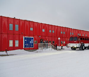 A large red bus is ready to leave Casey station with the old team on board. Some of the new team stand on the station porch waving goodbye to the people on the bus