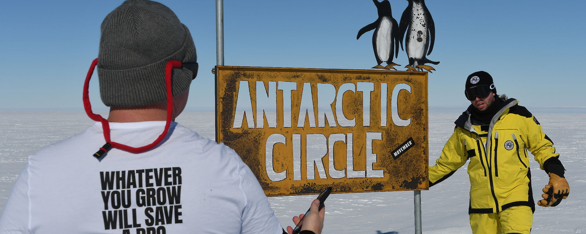 man's back wearing a movember t-shirt with another man in yellow survival clothing at the antarctic circle sign