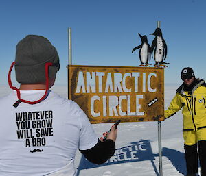 man's back wearing a movember t-shirt with another man in yellow survival clothing at the antarctic circle sign