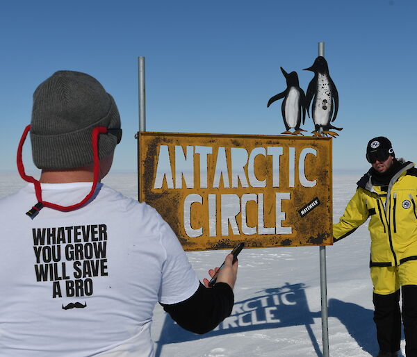 man's back wearing a movember t-shirt with another man in yellow survival clothing at the antarctic circle sign