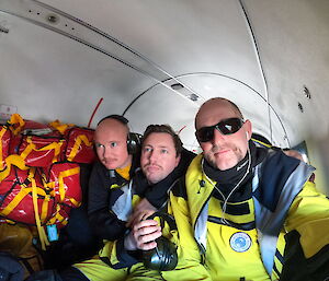 3 men in yellow survival clothing sitting inside a Basler aircraft with red and yellow bags in the background