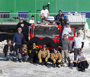 A group of people around a red Hagglund with a giant black wooden moustache in front of a green building