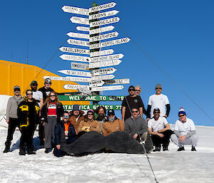 A group of people with the giant moustache in front of a group of sign posts and a yellow building on snow