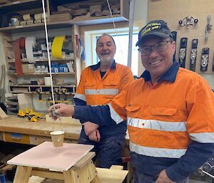 A man in a workshop stands beside a stool placed on a table.  A small pot of paint is suspended form a string above the stool.  Another man stands behind, both smiling to camera.