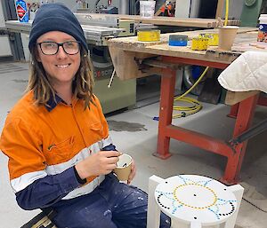 A man kneels on the ground next to an almost finished stool which he has been painting.