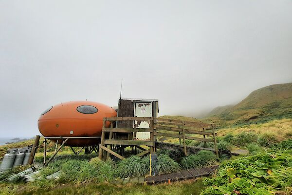 A red field hut sits amongst the green grass and grey clouds